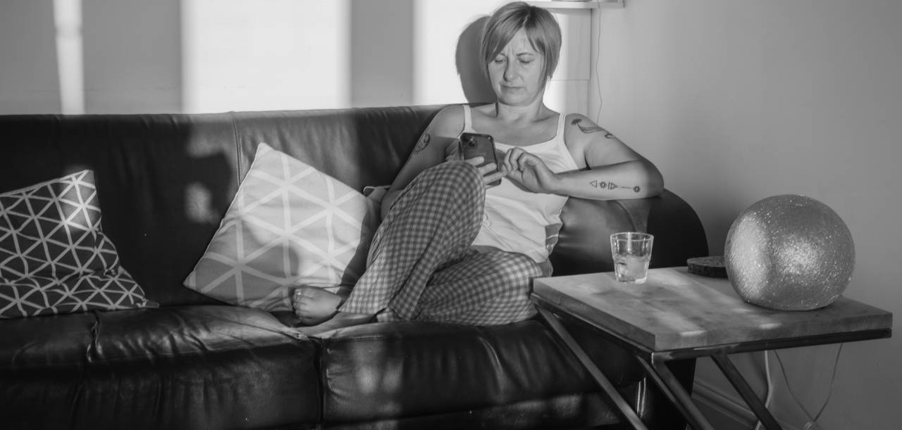 A black-and-white image of a woman sitting on her sofa in her living room wearing casual clothes and scrolling through her mobile phone.