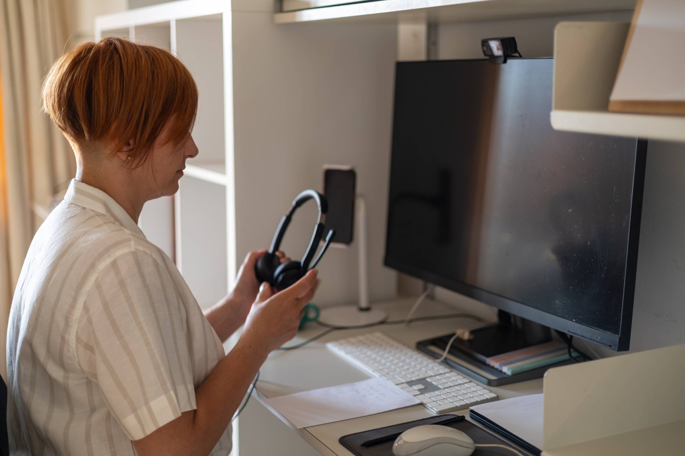 Woman sitting at her home office desk in front of a large screen holding a headset with microphone.