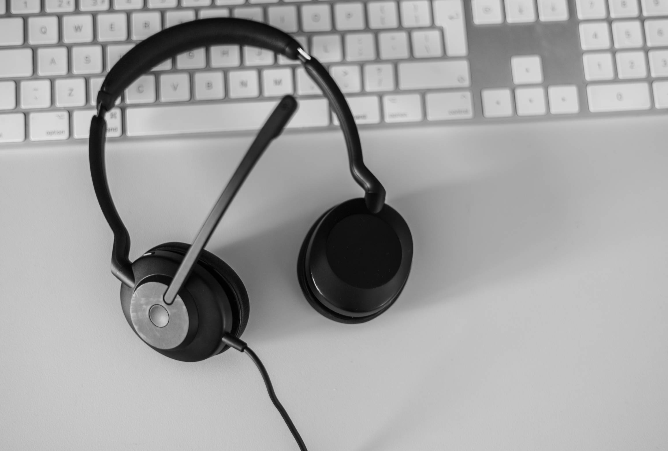 A black-and-white image of a headset with a microphone on a white desk with a white computer keyboard.