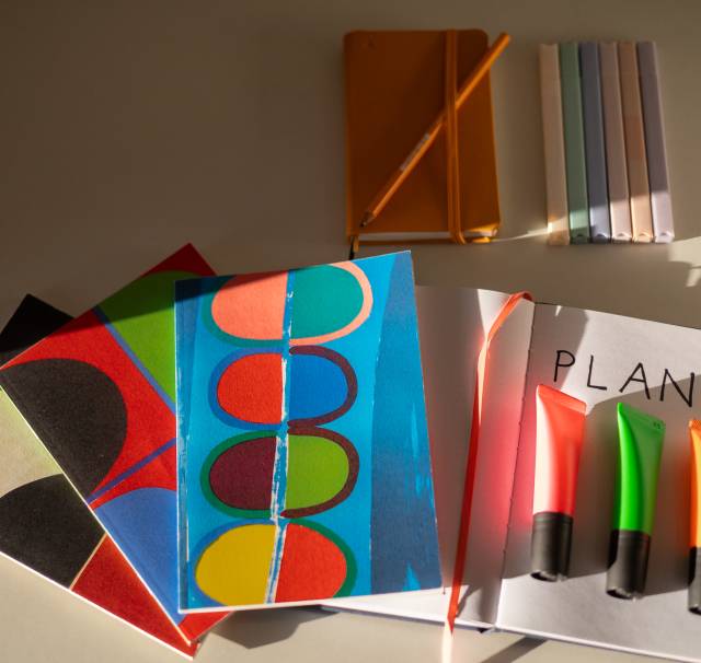 Colourful notebooks, pens and a sheet of paper with the writing Plan A (conference interpreter brief) arranged neatly on a white desk.