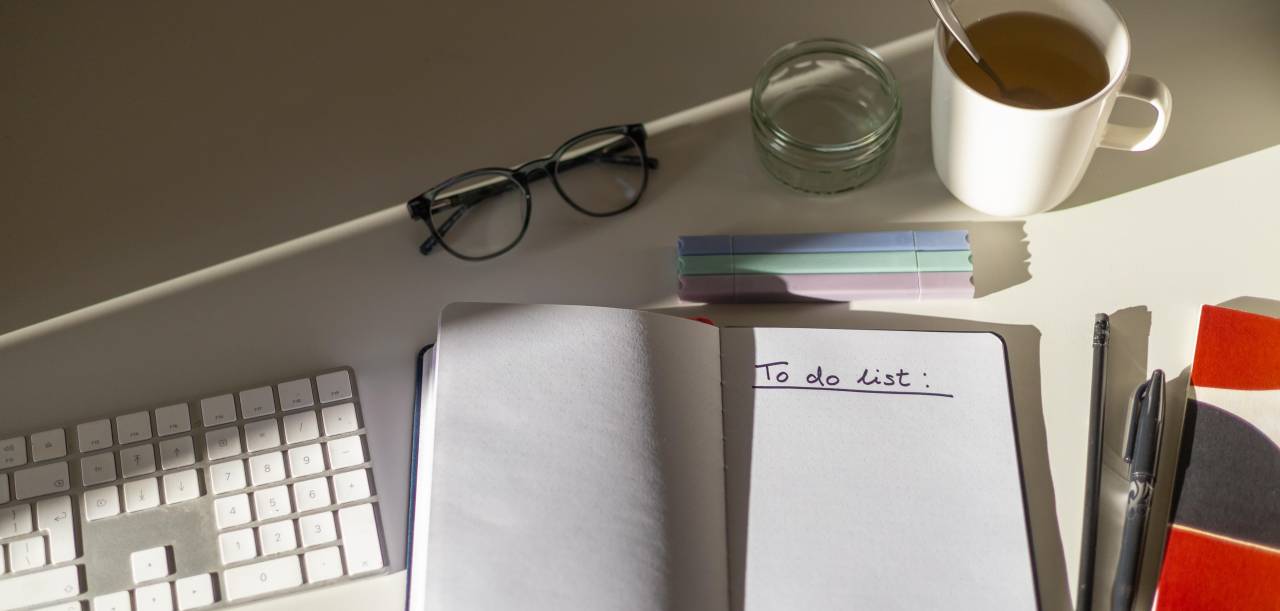 A conference interpreter home office desk featuring a keyboard, a notebook with a to-do-list, glasses and a tea mug.
